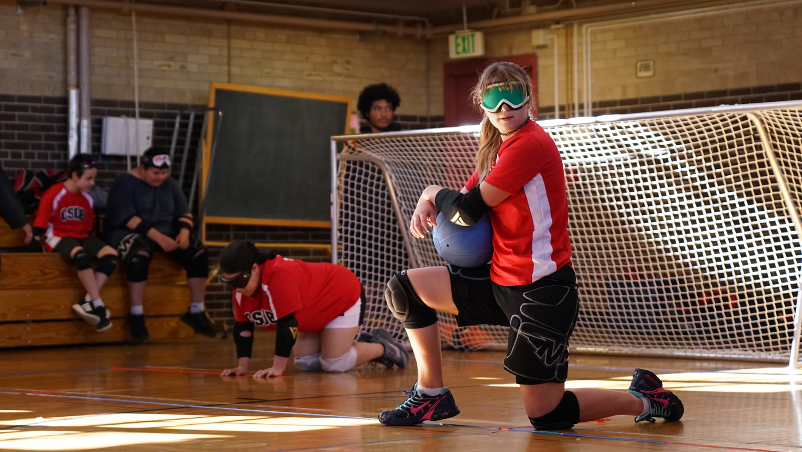 Tayler kneels on the court holding the goalball, teammates behind her.